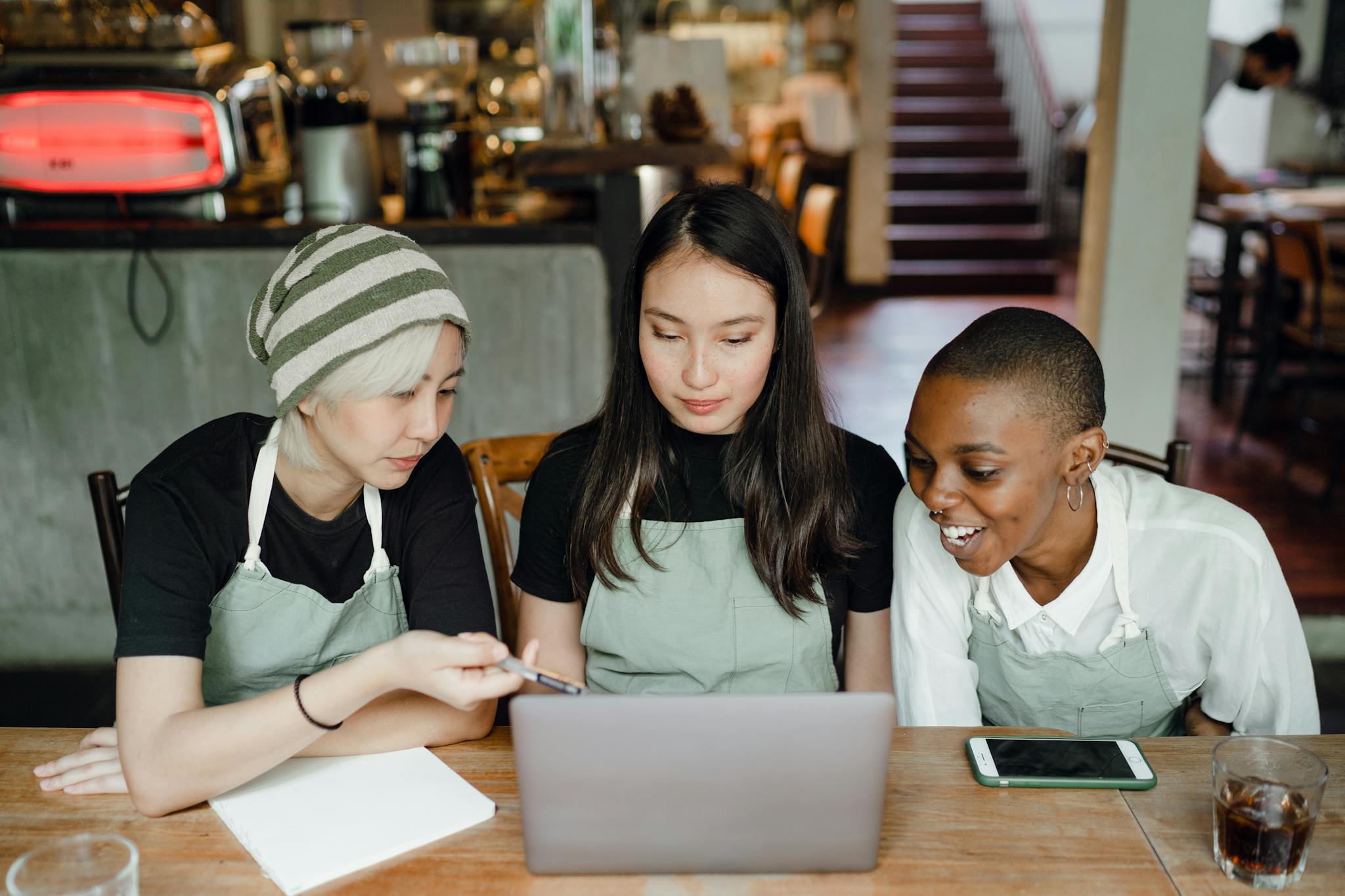 Three caf&eacute; workers in aprons collaborate cheerfully using a laptop indoors.